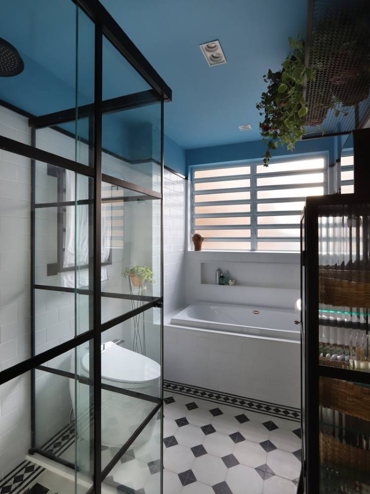Teto-Colorido-6 Esta é uma imagem de um banheiro moderno com um teto colorido azul. Possui uma banheira grande sob uma janela com persianas, e um box de chuveiro com estrutura preta e vidro. O piso apresenta um padrão quadriculado em branco e preto, e há plantas penduradas.