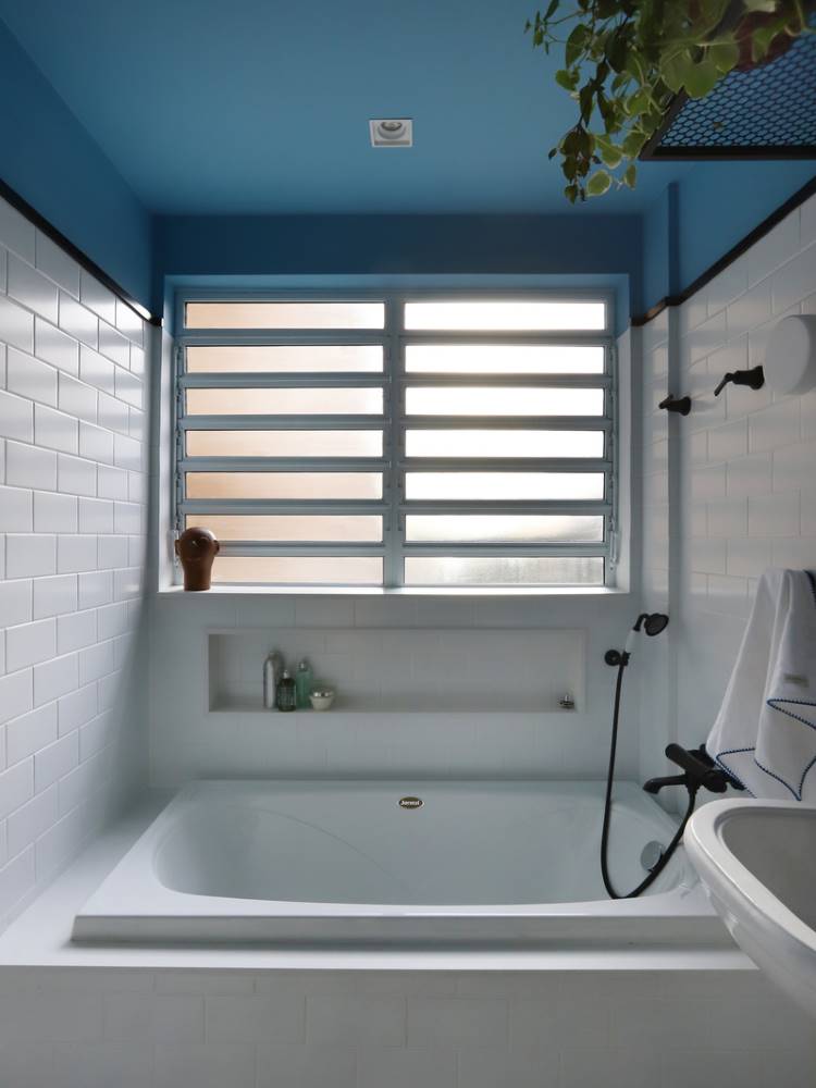 Teto-Colorido-7 Esta imagem exibe um banheiro com um teto colorido azul-claro. Uma banheira branca retangular com nicho embutido ocupa o centro, sob uma janela com persianas horizontais. As paredes são revestidas com azulejos brancos tipo metrô, e plantas penduradas complementam a decoração.