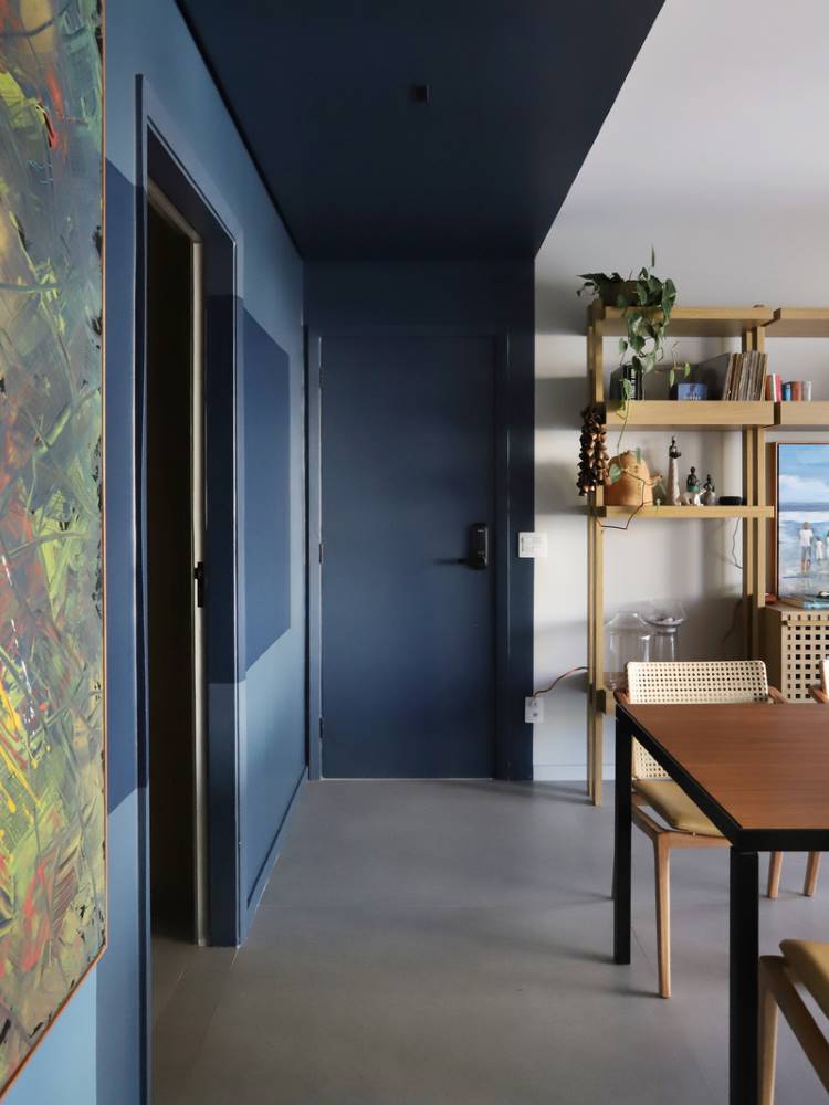 Teto-Colorido-8 Esta imagem mostra um corredor com paredes e um teto colorido em tons de azul escuro. Uma porta azul discreta se destaca. À direita, um ambiente com prateleiras de madeira, objetos decorativos e uma mesa de jantar sugere uma sala de estar. Uma obra de arte vibrante adiciona contraste.