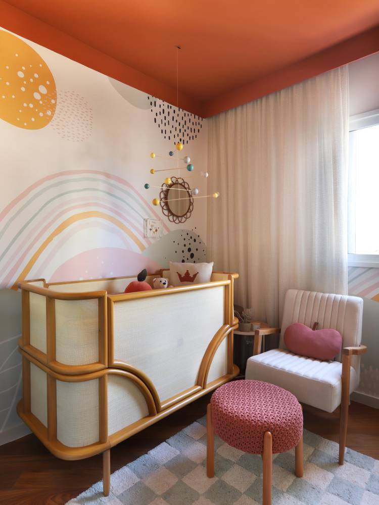 Teto-Colorido-10 Esta é uma imagem de um quarto de bebê vibrante com um teto colorido laranja. O berço de madeira clara com tela de vime se destaca contra a parede com um mural de arco-íris e bolinhas. Um pufe rosa adiciona um toque de cor, completando o ambiente lúdico.
