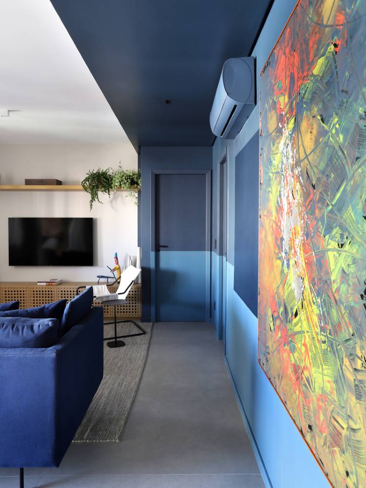 Teto-Colorido-9 Esta imagem mostra uma sala de estar moderna com um sofá azul e uma grande TV. Um corredor adjacente exibe paredes azuis com um teto colorido em azul-marinho. Uma porta discreta se mistura à parede, e uma obra de arte abstrata vibrante adiciona um toque de cor.