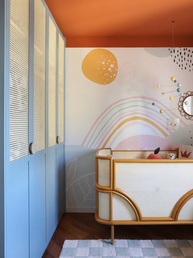 Teto-Colorido-11 Esta imagem mostra um quarto de bebê com um teto colorido laranja vibrante. Há um berço de madeira clara com tela de vime e um mural de parede com arco-íris e formas abstratas. Um guarda-roupa azul com portas de vime adiciona um toque de charme único ao ambiente.