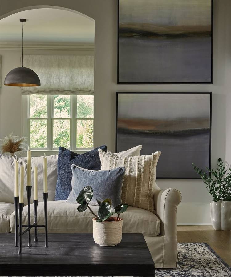 Almofadas-Para-Sofa-11 Uma sala de estar elegante apresenta um sofá claro com almofadas azuis e creme. Uma mesa de centro escura exibe um vaso de plantas e um castiçal. Duas obras de arte abstratas em tons suaves adornam a parede cinza, e uma luminária pendente complementa o ambiente.