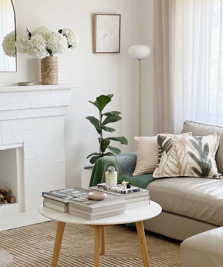 Almofadas-Para-Sofa-12 Uma sala de estar clara com sofá bege, almofadas estampadas e manta verde. Uma mesa de centro redonda exibe livros e decoração. Ao fundo, uma lareira branca, vaso com flores brancas e um espelho. Uma planta alta e uma luminária de piso completam o ambiente.