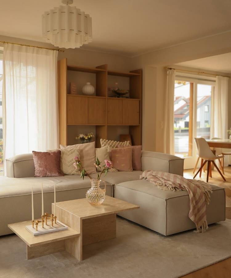 Almofadas-Para-Sofa-24 Uma sala de estar espaçosa com sofá em L de tom neutro, muitas almofadas e uma manta. Mesas de centro geométricas com velas e flores. Uma estante de madeira clara na parede, cortinas leves e grandes janelas trazem luminosidade. O ambiente é acolhedor e elegante.