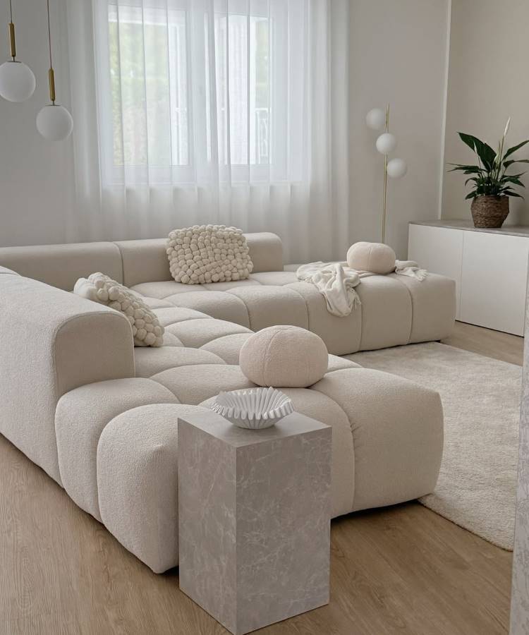 Almofadas-Para-Sofa-18 Uma sala de estar minimalista com sofá modular bege claro e almofadas para sala texturizadas e redondas. Uma mesa lateral quadrada de mármore claro exibe um bowl branco. Cortinas brancas translúcidas e uma luminária de piso completam o ambiente suave e sereno, com um toque de verde de uma planta.