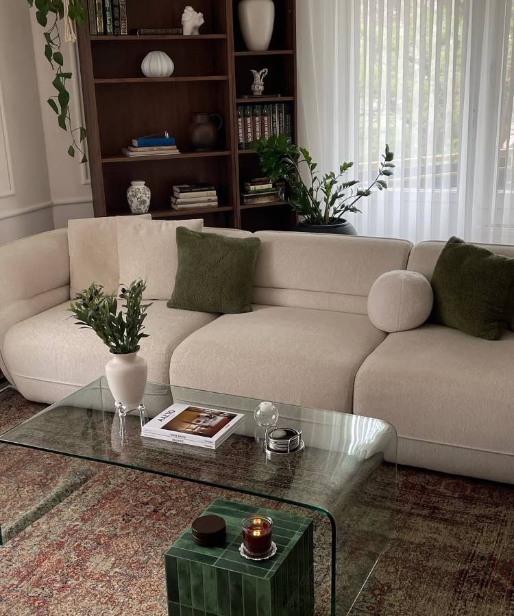 Almofadas-Para-Sofa-22 Uma sala de estar elegante com sofá bege, almofadas para sala verdes e redondas. Uma mesa de centro de vidro curvado exibe um vaso com folhagens e livros. Ao fundo, uma estante escura com livros e objetos decorativos. Cortinas brancas e plantas penduradas completam o ambiente.