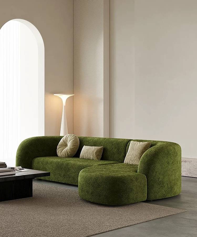 Almofadas-Para-Sofa-5 Uma sala minimalista e moderna com um sofá verde musgo curvado e um pufe combinando. As almofadas para sala em tons neutros complementam o estofamento texturizado. Ao fundo, um arco elegante e uma luminária de piso escultural. O ambiente exala design contemporâneo e sofisticação.