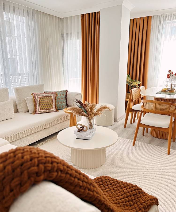 Almofadas-Para-Sofa-16 Uma sala de estar e jantar integrada, com sofá creme e almofadas para sala estampadas. A mesa de centro redonda exibe um arranjo floral. Cortinas em tons terrosos complementam a área de jantar com mesa branca e cadeiras de madeira. O ambiente é convidativo e harmonioso.