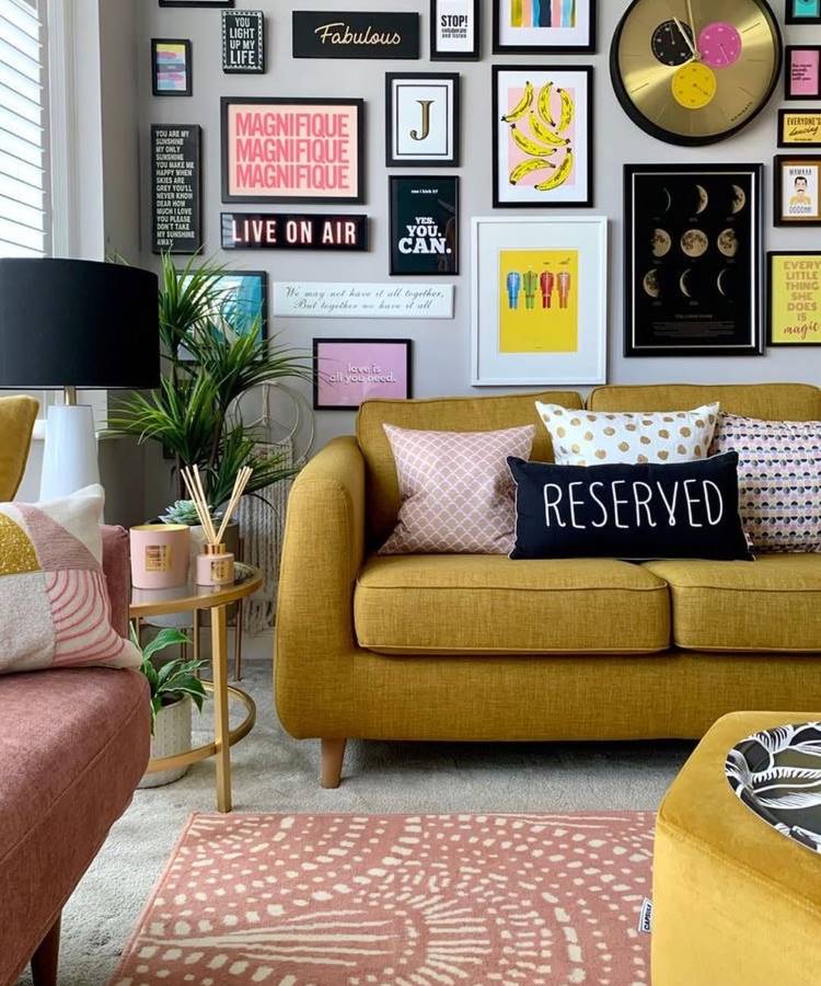 Almofadas-Para-Sofa-2-1-1 Uma sala vibrante com sofá mostarda e almofadas para sala decorativas, incluindo uma com a palavra "RESERVED". A parede cinza é coberta por uma galeria de quadros coloridos e artísticos. Um tapete rosa com estampa geométrica e uma mesa lateral dourada completam o estilo eclético e divertido.
