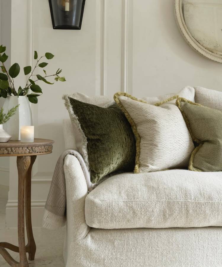 Almofadas-Para-Sofa-10 Uma sala elegante com sofá claro e almofadas para sala em tons de verde e creme, com texturas e franjas. Ao lado, uma mesa de madeira redonda exibe um vaso com folhagens e uma vela acesa. Um espelho redondo na parede e uma luminária preta completam o ambiente sofisticado.