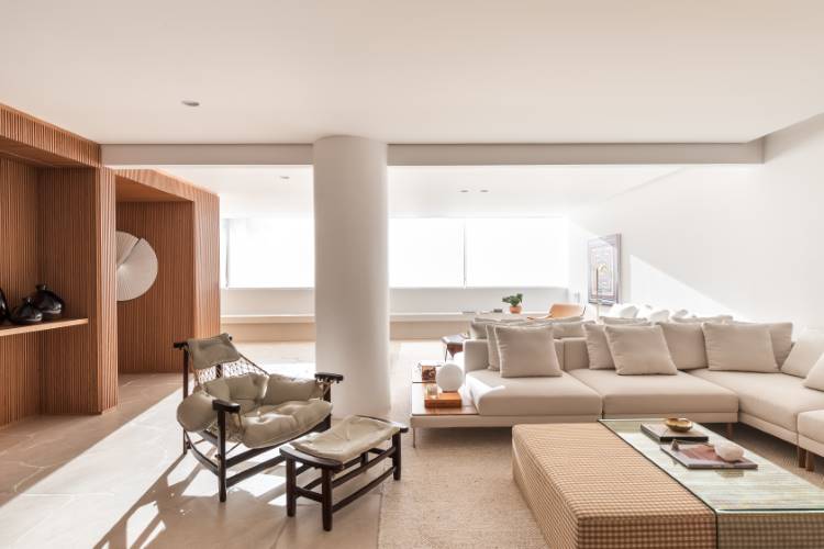 Apartamento-Reforma-Compelta-6 A imagem mostra uma sala de estar moderna e espaçosa. Há um sofá modular bege, uma poltrona de madeira clara com estofamento bege e uma mesa de centro com tampo de vidro. Uma parede ripada de madeira adiciona textura, e grandes janelas iluminam o ambiente.