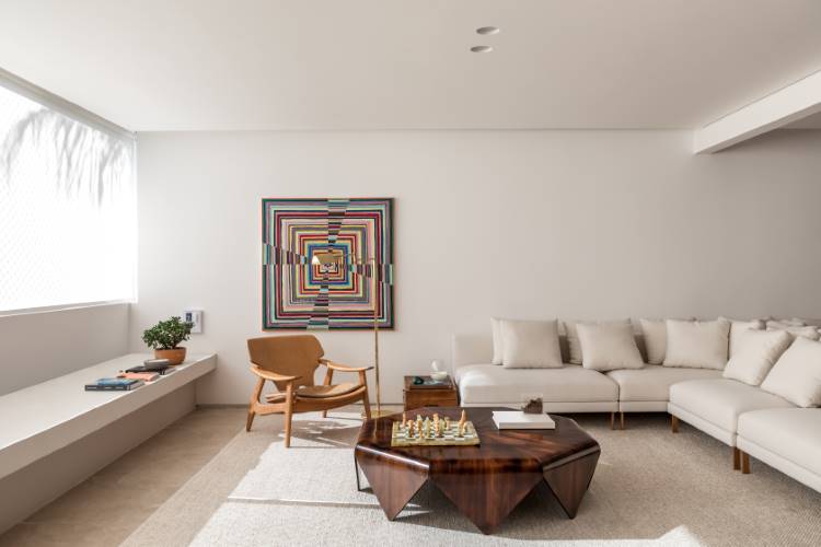 A imagem apresenta uma sala de estar moderna e iluminada, com um sofá branco e uma poltrona de madeira clara. Uma mesa de centro hexagonal de madeira escura e uma obra de arte colorida na parede centralizam a atenção, complementadas por uma ampla janela que inunda o espaço com luz natural.