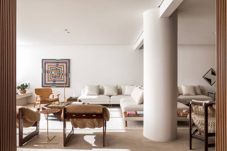 A imagem apresenta uma sala de estar moderna e iluminada, com um sofá branco e uma poltrona de madeira clara. Uma mesa de centro hexagonal de madeira escura e uma obra de arte colorida na parede centralizam a atenção, complementadas por uma ampla janela que inunda o espaço com luz natural.