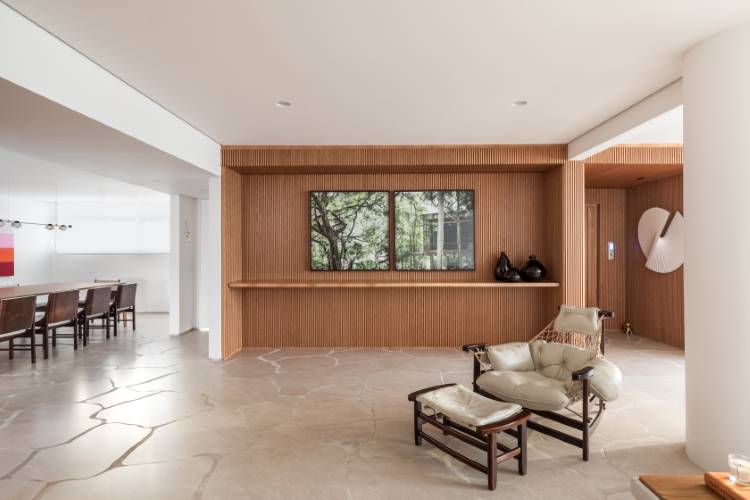 A imagem exibe um interior sofisticado com uma parede ripada de madeira, que serve de fundo para duas obras de arte de paisagem. Uma poltrona de madeira escura com estofamento claro e um pufe estão em primeiro plano. Ao fundo, uma sala de jantar com mesa e cadeiras de madeira escura é visível.