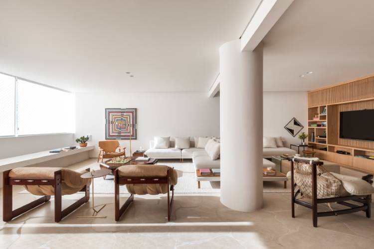 A imagem apresenta uma sala de estar moderna e iluminada, com um sofá branco e uma poltrona de madeira clara. Uma mesa de centro hexagonal de madeira escura e uma obra de arte colorida na parede centralizam a atenção, complementadas por uma ampla janela que inunda o espaço com luz natural.