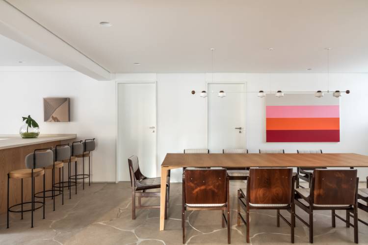 Apartamento-Reforma-Compelta-9 A imagem mostra uma sala de jantar moderna e minimalista. Uma longa mesa de madeira com cadeiras escuras ocupa o centro, sob uma luminária contemporânea. Uma obra de arte colorida decora a parede, e um balcão com bancos altos à esquerda completa o espaço.
