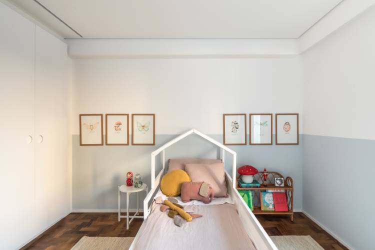 Apartamento-Reforma-Compelta-10 A imagem mostra um quarto infantil aconchegante com uma cama em formato de casinha e roupa de cama rosa. As paredes são brancas na parte superior e cinza-claro na inferior, decoradas com quadros de desenhos. Um armário branco e uma estante com livros e brinquedos completam o ambiente lúdico.