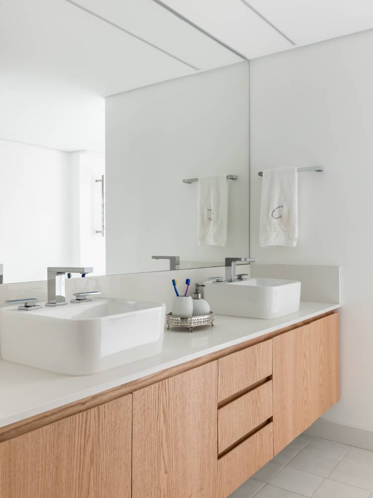 Um banheiro moderno e claro com duas pias retangulares brancas sobre um balcão de madeira clara. Um grande espelho reflete o ambiente, com toalhas brancas penduradas. O espaço é limpo e funcional, com torneiras prateadas e detalhes discretos.
