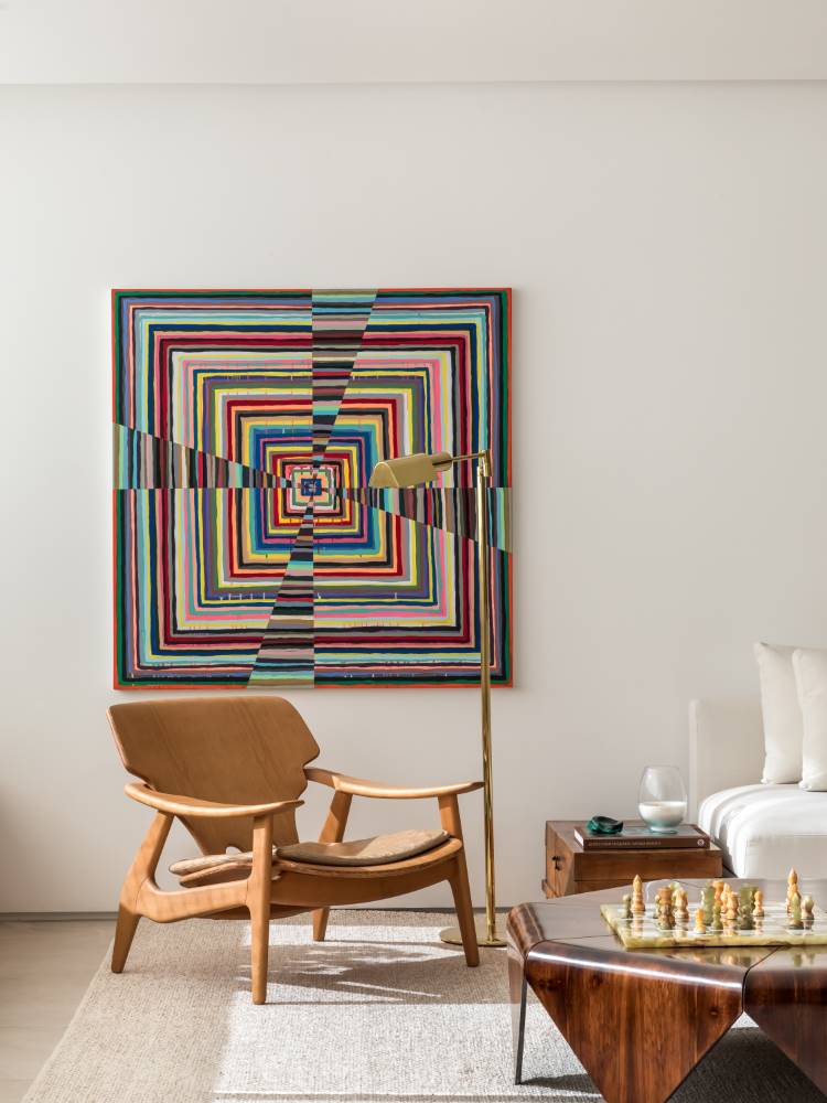Uma sala de estar elegante com uma poltrona de madeira clara e estofamento discreto. Atrás, uma grande obra de arte abstrata colorida adiciona um vibrante ponto focal. Uma mesa de centro de madeira escura com um tabuleiro de xadrez está em primeiro plano, complementando o ambiente sofisticado.