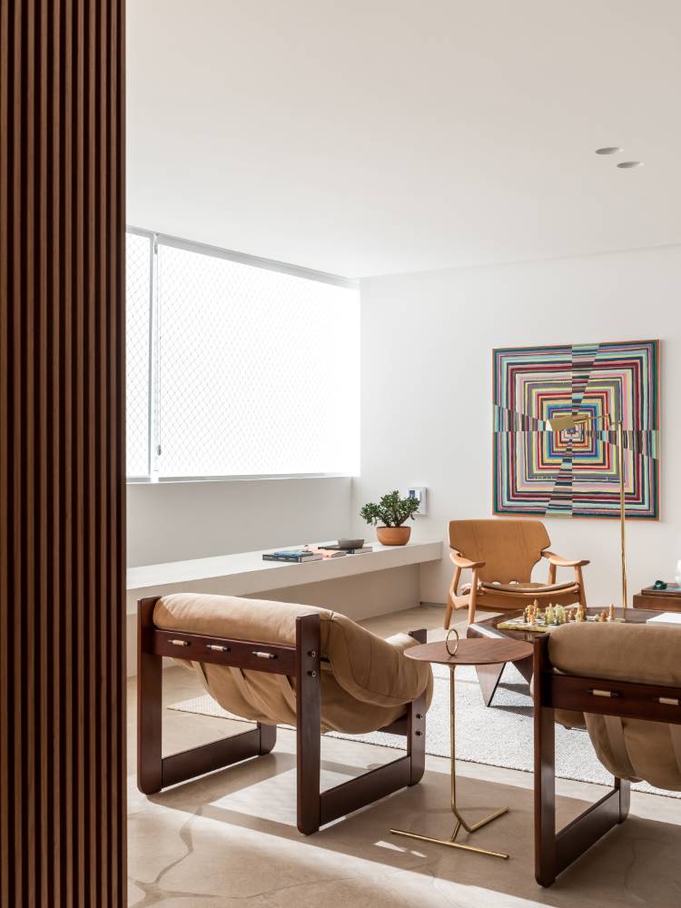 A imagem exibe uma sala de estar moderna e iluminada, com duas poltronas estofadas em tom bege em primeiro plano. Ao fundo, uma poltrona de madeira e uma vibrante obra de arte abstrata adornam a parede. Uma parede ripada de madeira à esquerda adiciona textura, e uma janela longa ilumina o espaço.