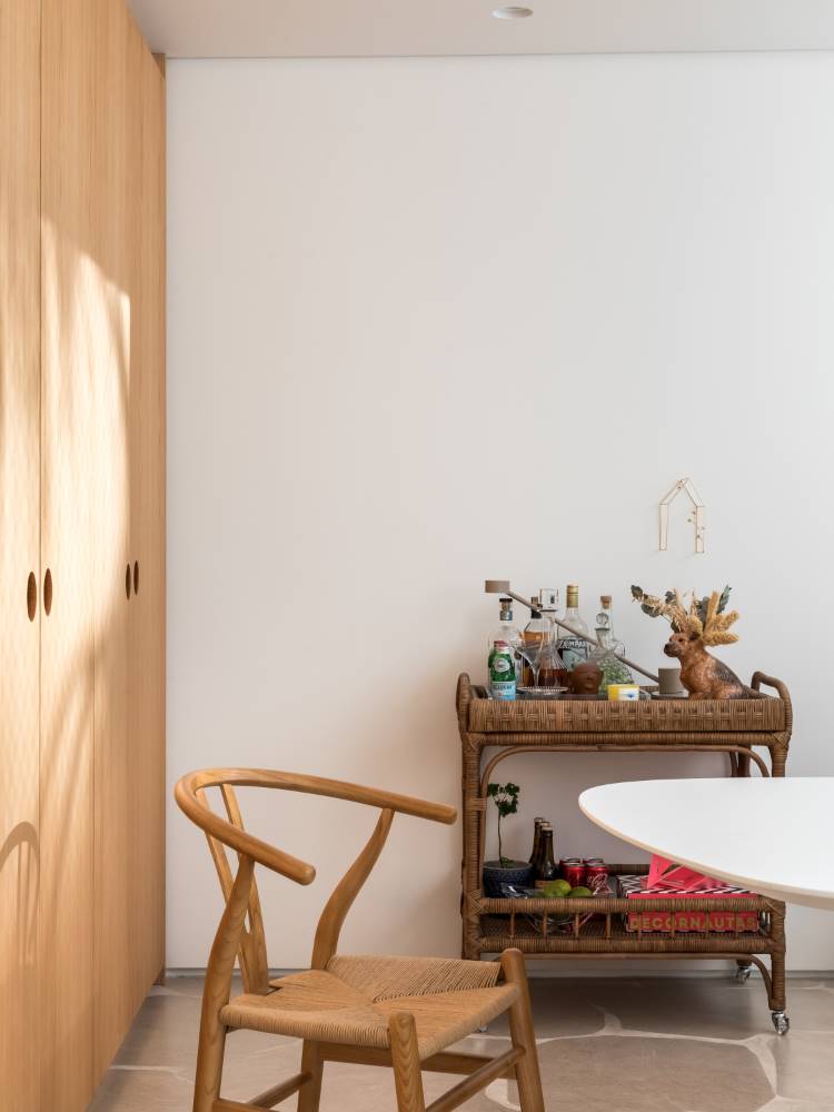 A imagem mostra um canto de sala com um carrinho de bebidas de vime, decorado com garrafas e objetos, incluindo uma rena. Uma cadeira de madeira com assento de palha está em primeiro plano, e armários de madeira clara com puxadores escuros ocupam a parede esquerda.