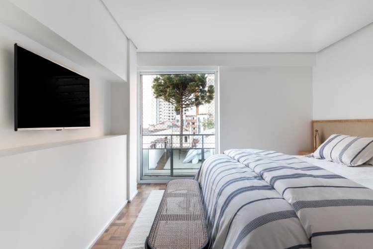 Apartamento-Reforma-Compelta-11 Um quarto moderno e minimalista com uma cama de madeira clara e roupa de cama listrada em azul e branco. Uma grande janela oferece vista para árvores e edifícios. Uma televisão de tela plana está montada na parede oposta, e um banco de vime repousa aos pés da cama.