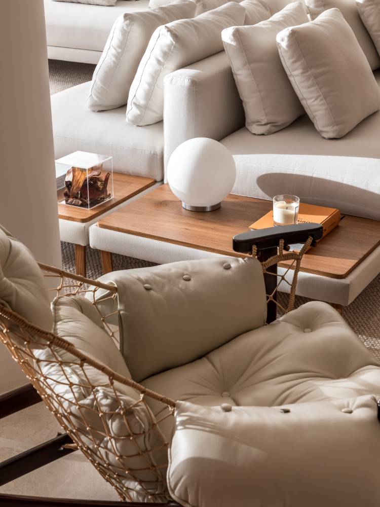 Uma sala de estar aconchegante com um sofá modular bege e uma poltrona de vime com almofadas claras em primeiro plano. Mesas laterais de madeira clara com uma luminária esférica e objetos decorativos estão dispostas ao lado do sofá.