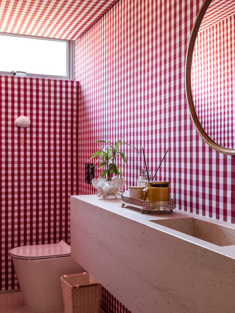 Um banheiro vibrante com paredes e teto cobertos por um papel de parede de xadrez vermelho e branco. Uma pia de pedra clara com um espelho redondo dourado acima. Um vaso de planta e acessórios decoram a bancada, e um vaso sanitário discreto está à esquerda.