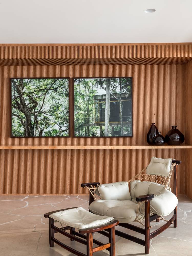 Um ambiente elegante com uma parede ripada de madeira que emoldura duas obras de arte com paisagens arbóreas. Em primeiro plano, uma poltrona de madeira escura e vime com estofamento claro e um pufe convidam ao relaxamento. Vasos decorativos escuros adornam a prateleira.