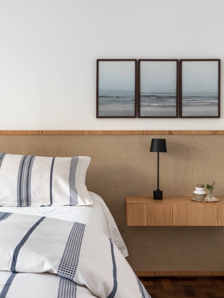 Um quarto sereno com uma cama coberta por roupa de cama listrada em azul e branco. A cabeceira da cama é de um material natural com textura. Acima da cama, três quadros exibem paisagens marinhas em tons suaves. Uma mesa de cabeceira flutuante de madeira clara com uma luminária preta completa o ambiente.