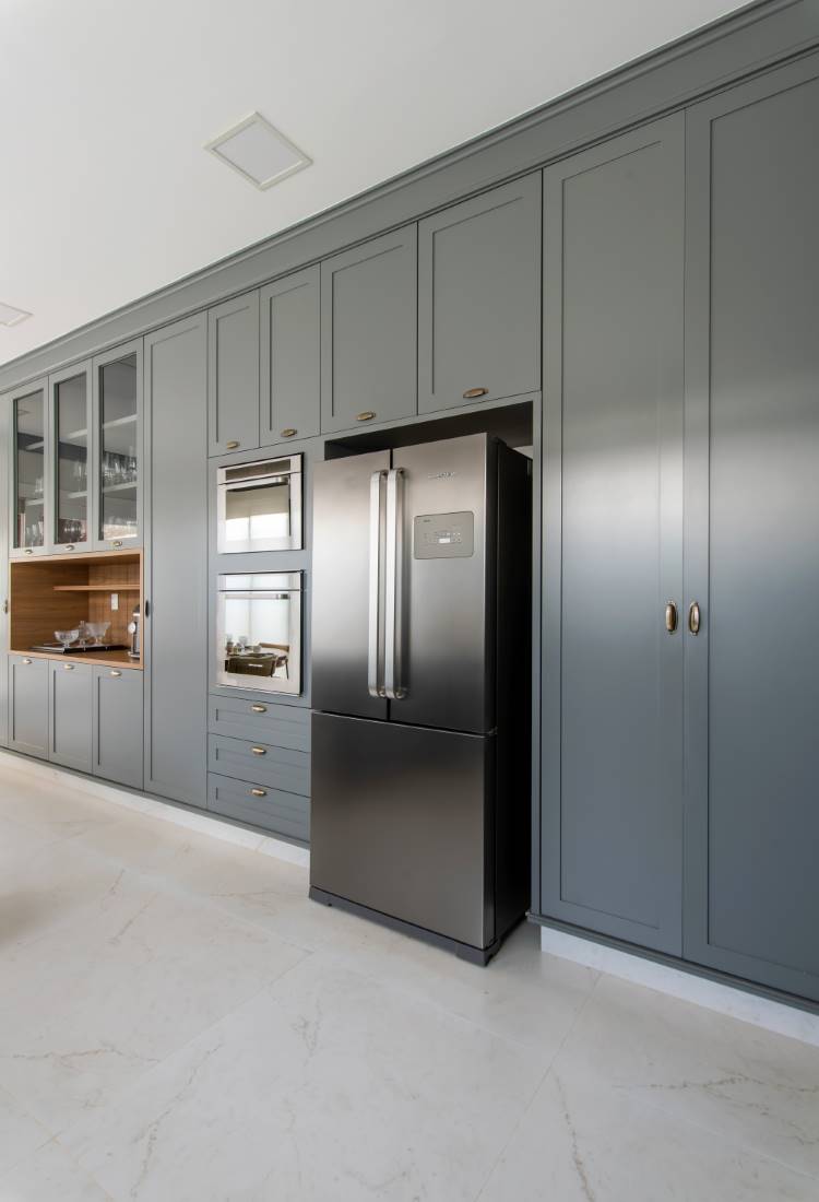 Cozinha-Com-Marcenaria-Planejada-8 Uma cozinha moderna com armários cinza que vão do chão ao teto, alguns com portas de vidro. Há uma geladeira de aço inoxidável e fornos embutidos. O piso é de mármore branco, e a iluminação é embutida.
