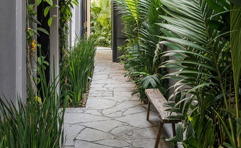 Corredor externo com banco de madeira compacto ao lado de paisagismo com plantas tropicais e caminho de pedras claras.