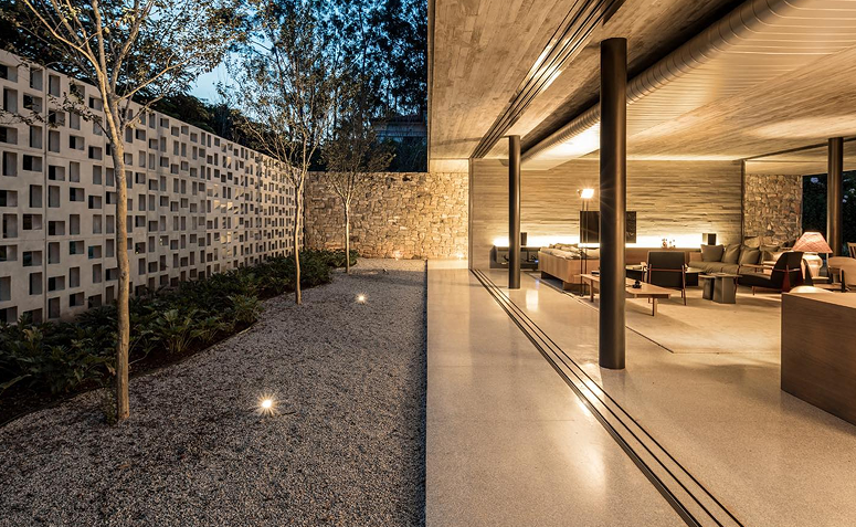 Corredor externo com iluminação no piso destacando o caminho de pedriscos e o jardim ao lado de uma casa contemporânea.