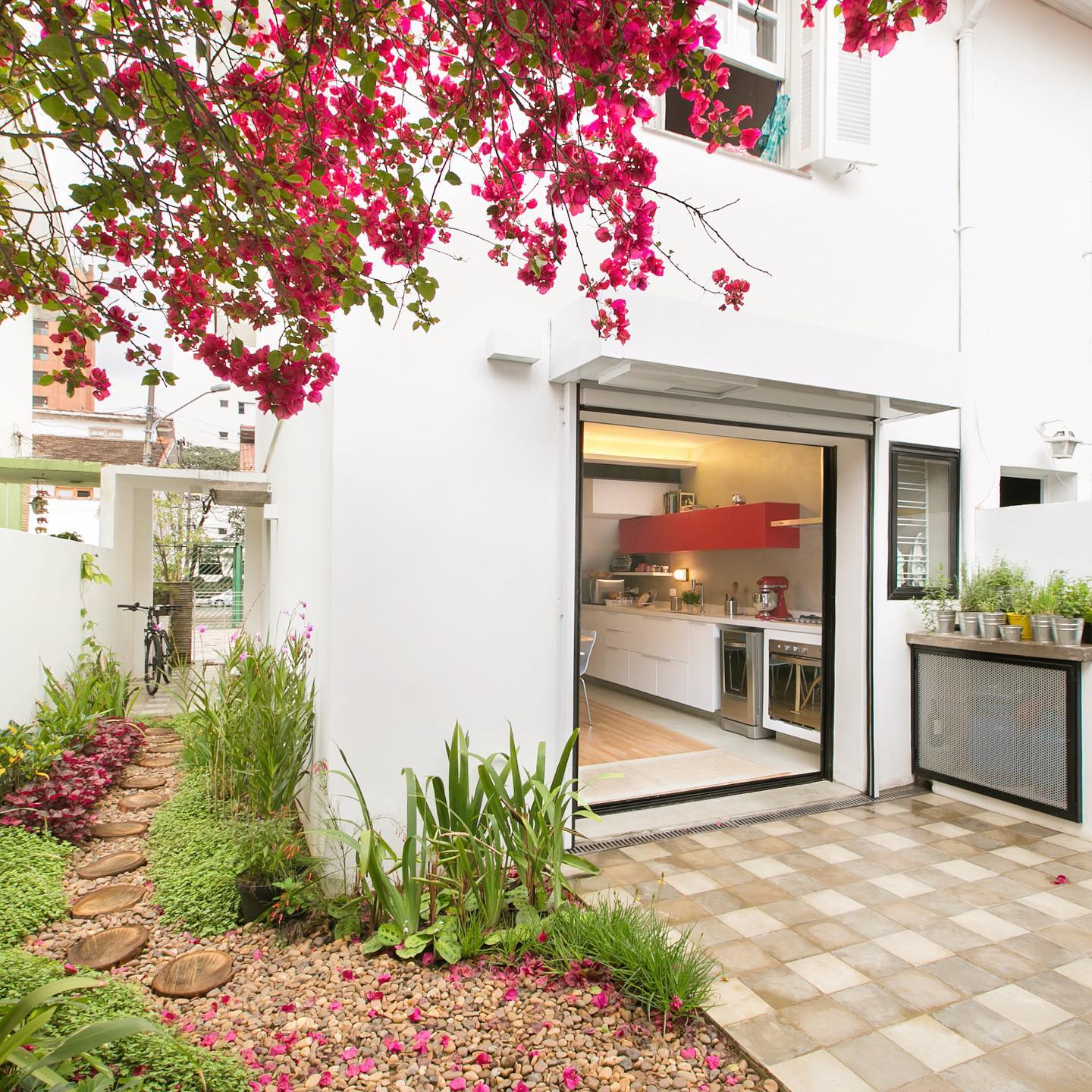 Corredor externo com pisantes de madeira natural formando caminho entre plantas tropicais e paisagismo lateral.