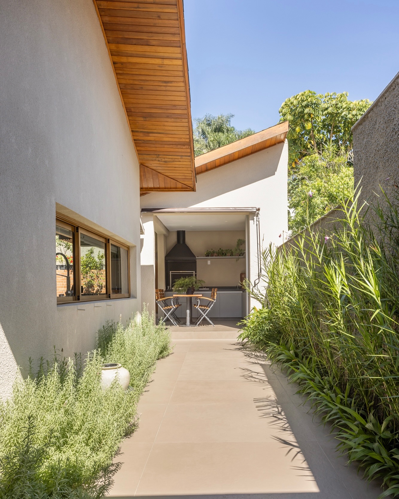 Passagem lateral com piso claro, paisagismo com plantas ornamentais e área gourmet com churrasqueira ao fundo.
