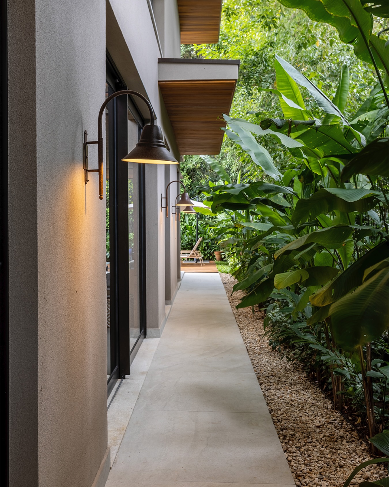 Corredor externo com iluminação em arandelas na parede e paisagismo tropical ao lado do caminho.