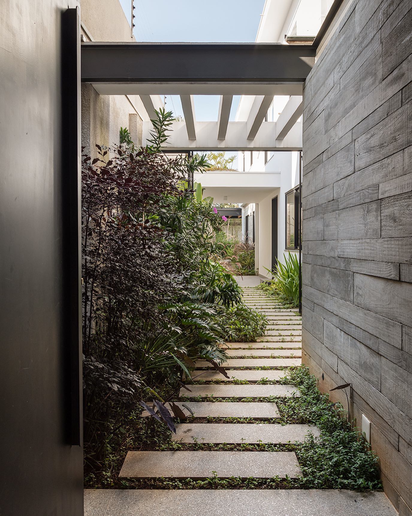 Corredor externo com pisantes de concreto intercalados com vegetação e jardim lateral.