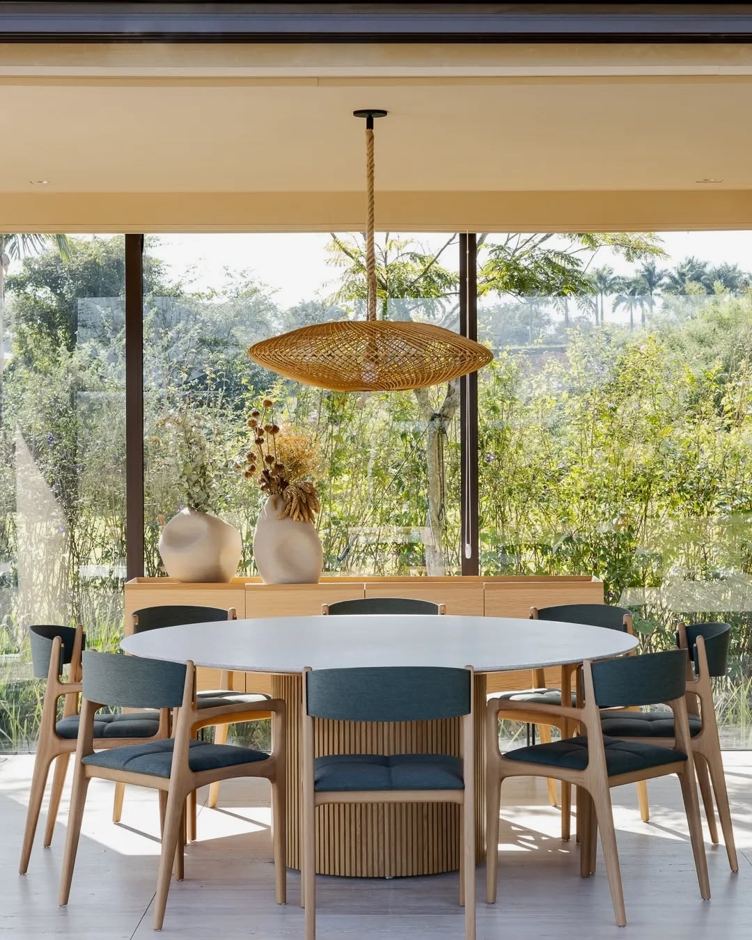 Sala de jantar com mesa redonda, cadeiras de madeira estofadas e luminária de fibra natural diante de grandes janelas
