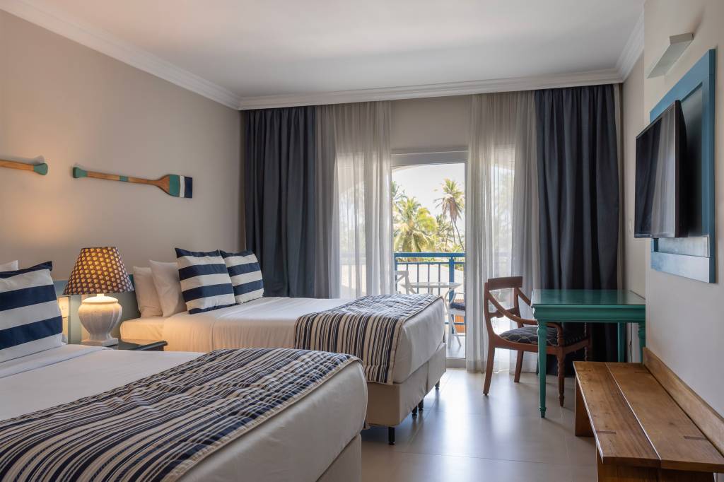 Quarto de hotel com duas camas, com roupa de cama branca e azul. Há dois remos decorativos na parede atrás da cama. Há um banco longo e uma TV à frente das camas. No fundo do quarto, fica uma porta de vidro, com cortinas, que dá para uma sacada com vista para a piscina.