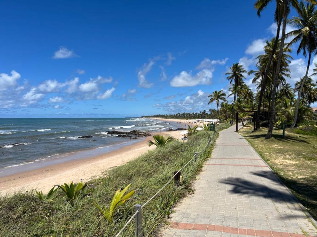 Calçadão à beira da praia na Costa do Sauípe, ladeado por vegetação e árvores.