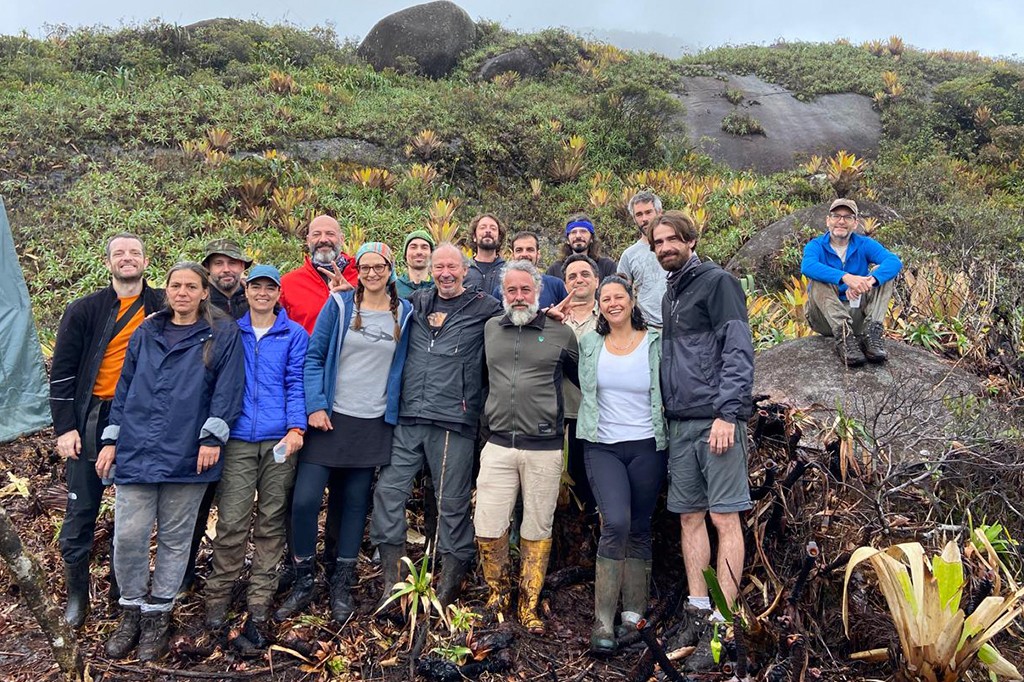 Grupo de 18 pessoas, incluindo homens e mulheres de diversas idades, sorrindo e posando para foto em trilha montanhosa. Vestem roupas e botas de trekking, alguns com capas de chuva. Um homem de azul está sentado em uma rocha ao fundo. O cenário é de vegetação rasteira e rochas, com céu nublado
