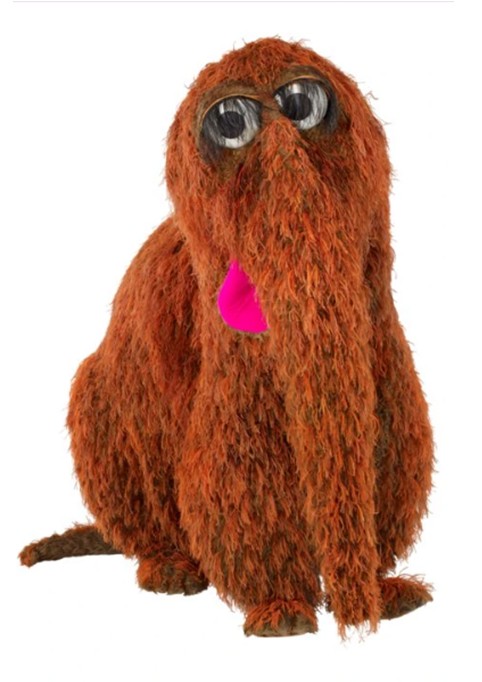 Boneco Snuffy, personagem do programa Vila Sésamo, sentado, com corpo coberto por pelos marrons, olhos grandes e redondos, e uma língua rosa vibrante saindo da boca, sobre fundo branco