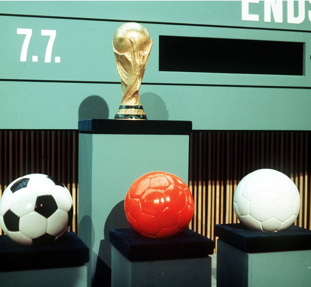A Taça da Copa do Mundo FIFA dourada em um pedestal preto, com três bolas de futebol em pedestais menores: uma branca e preta, uma laranja e uma branca. Ao fundo, um painel verde com a data