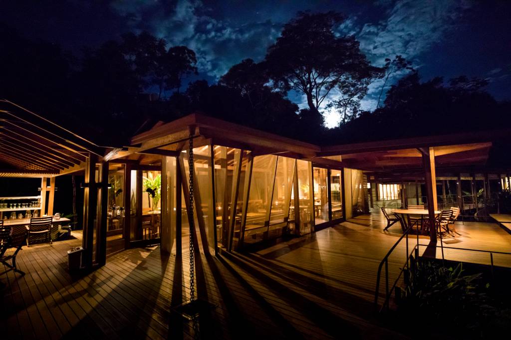 Um restaurante moderno com paredes de vidro e estrutura de madeira, iluminado por dentro, com mesas e cadeiras no deck de madeira ao ar livre, sob um céu noturno azul escuro com nuvens e árvores ao fundo