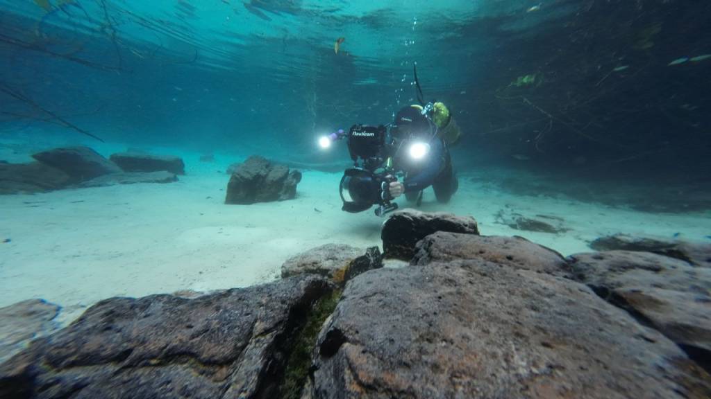 Mergulhador com equipamento de filmagem subaquática, incluindo câmera e luzes potentes, flutuando sobre um leito de areia clara com rochas escuras no fundo de um rio ou lago de águas cristalinas e azul-turquesa