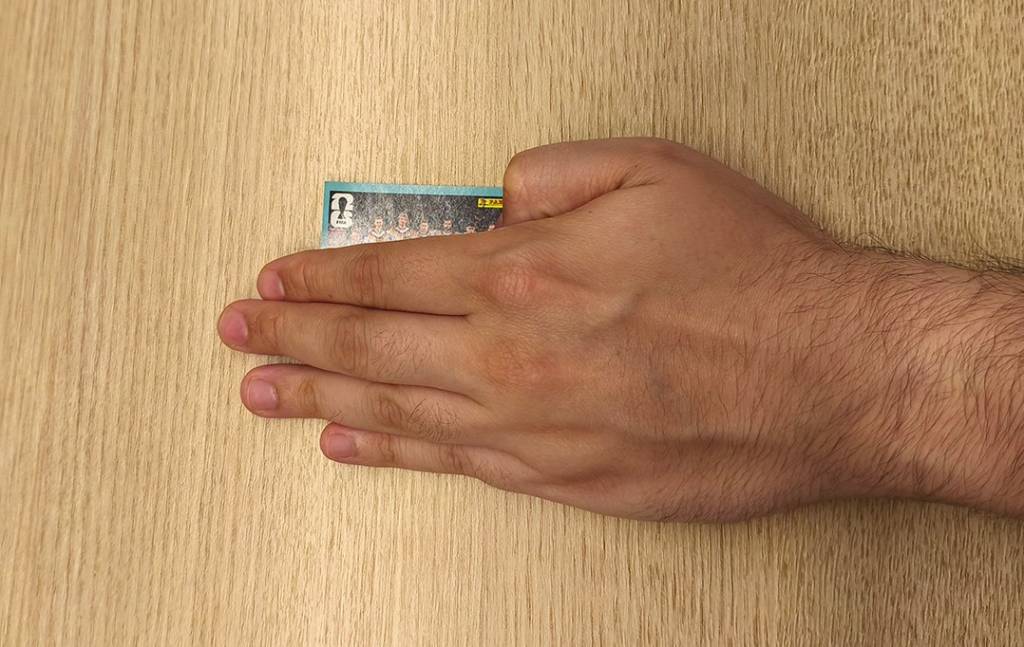 Mão masculina clara, com pelos no pulso, segurando um cromo de figurinhas da Panini com a imagem de um time de futebol, sobre uma superfície de madeira clara