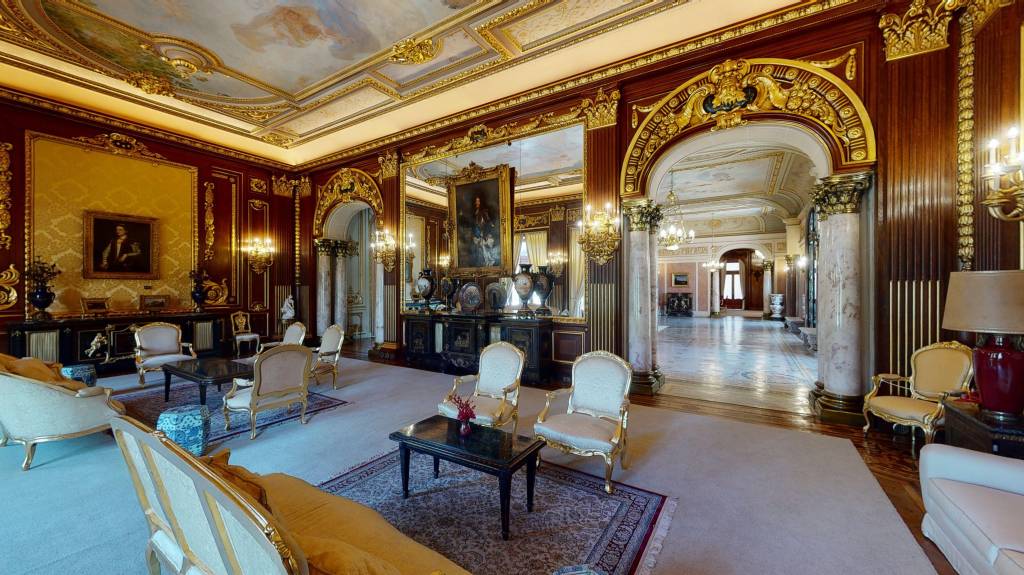 Salão luxuoso com paredes de madeira escura e detalhes dourados, teto pintado, espelhos grandes, sofás e poltronas clássicas com estofamento claro, mesas de centro escuras, tapetes estampados e lustres ornamentados. Ao fundo, um arco dourado leva a um corredor com piso de mármore e colunas. Foto sob licença CC BY-SA 4.0