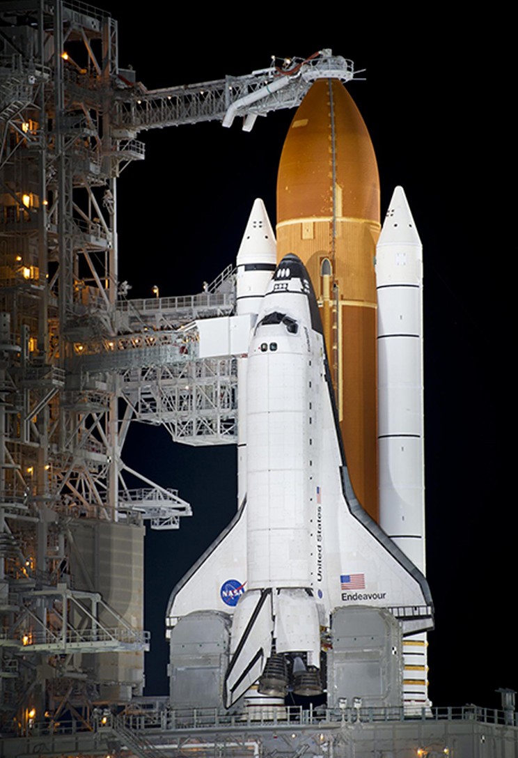 Ônibus espacial Endeavour branco acoplado a um tanque de combustível laranja e dois propulsores brancos, em uma plataforma de lançamento iluminada à noite