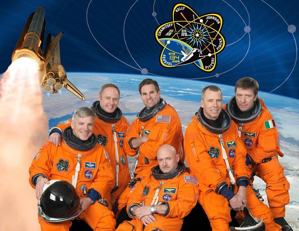 Seis astronautas sorridentes em trajes espaciais laranja posam com a Terra ao fundo, um ônibus espacial decolando e o emblema da missão STS-134